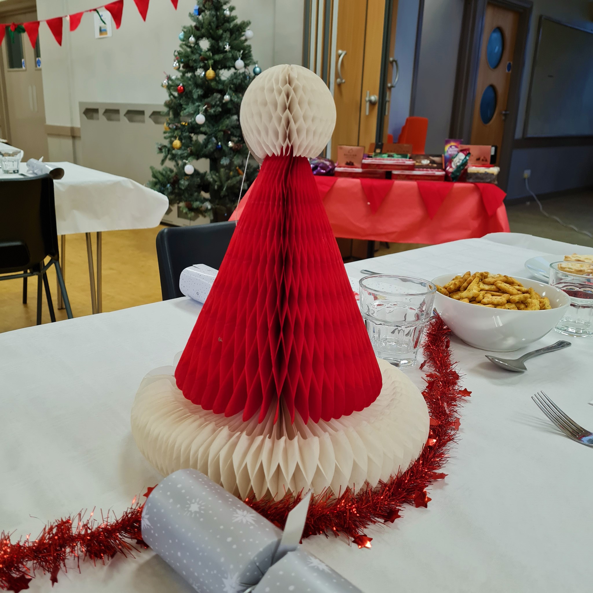 Christmas Santa hat decoration on decorated table before lunch. Christmas tree and gifts for guests in the background.