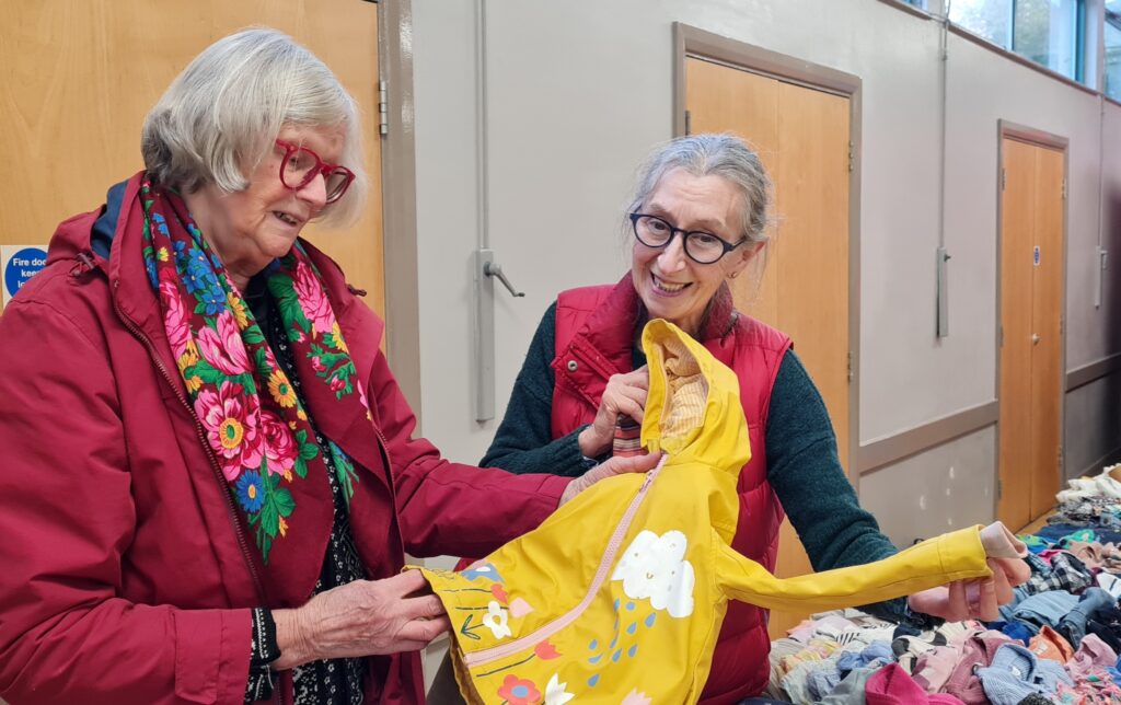 Two women with red jackets and grey hair hold up a yellow children's coat. They stand beside a table covered in clothes.