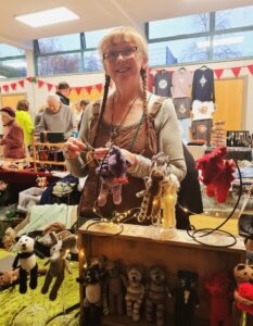 Woman with plaited hair knitting, standing beside a stall featuring a selection of knitted bears