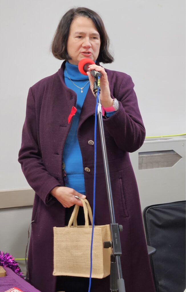 The Hon Catherine West MP wearing a purple coat and holding a beige bag, speaking into a microphone