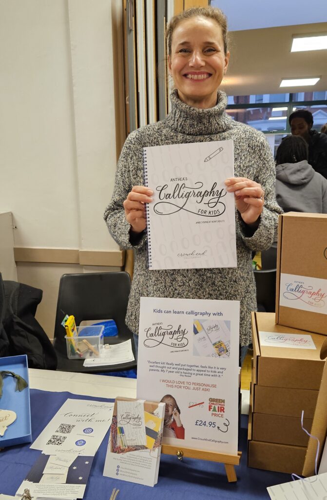 Woman in grey jumper smiling and standing up holding a piece of paper with calligraphy written on it in curling script